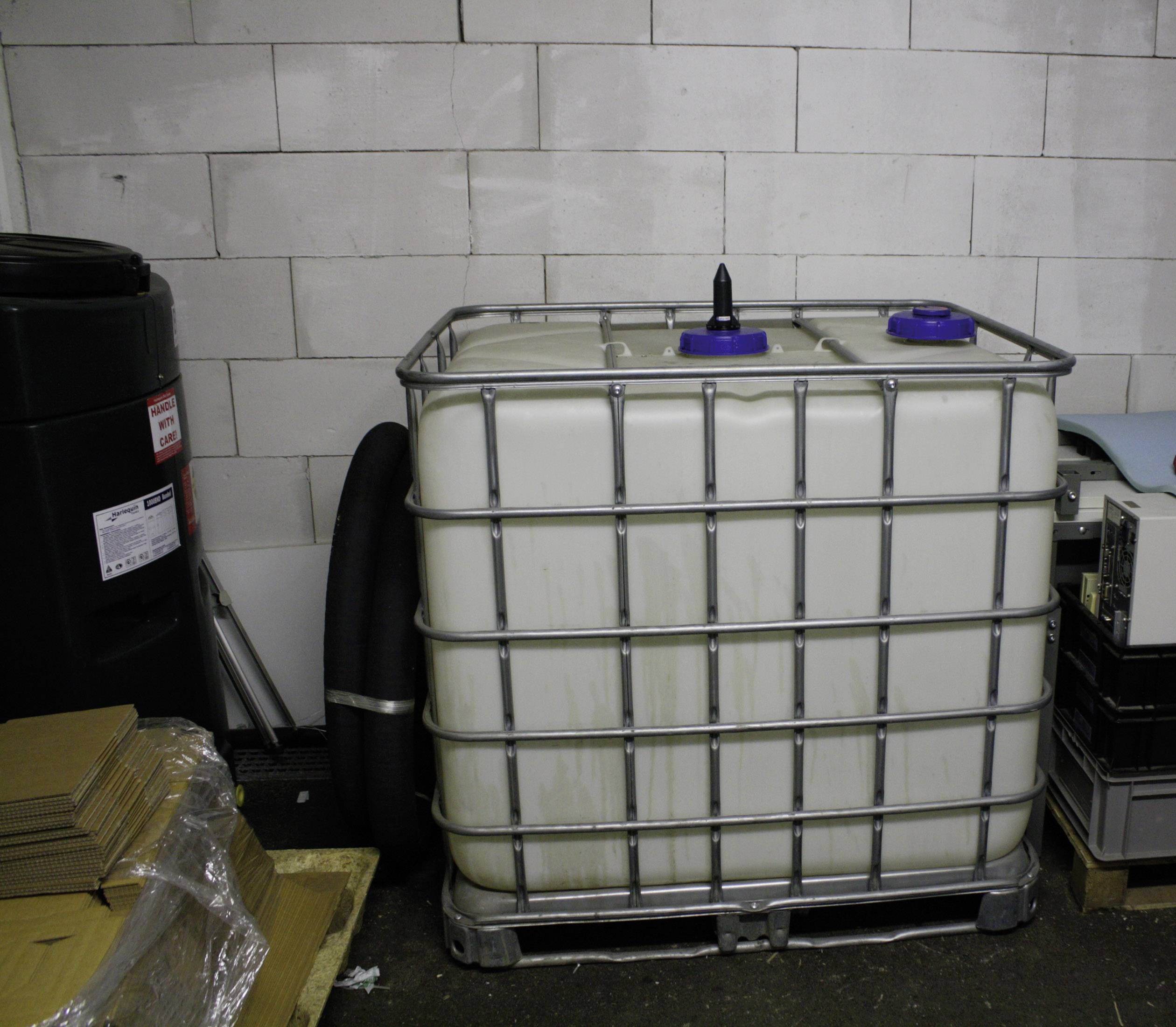 Ein großer weißer Kunststofftank in einem Metallrahmen steht in einem Lagerraum. Links daneben sind Kartons und ein schwarzer Zylinder.