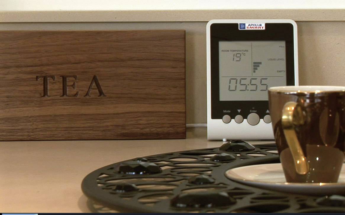 Eine braune Tasse auf einer Untertasse steht auf einem Tisch. Daneben ein Holzbehälter mit der Aufschrift 'TEA' und ein Thermometer mit zugehörigem Display.