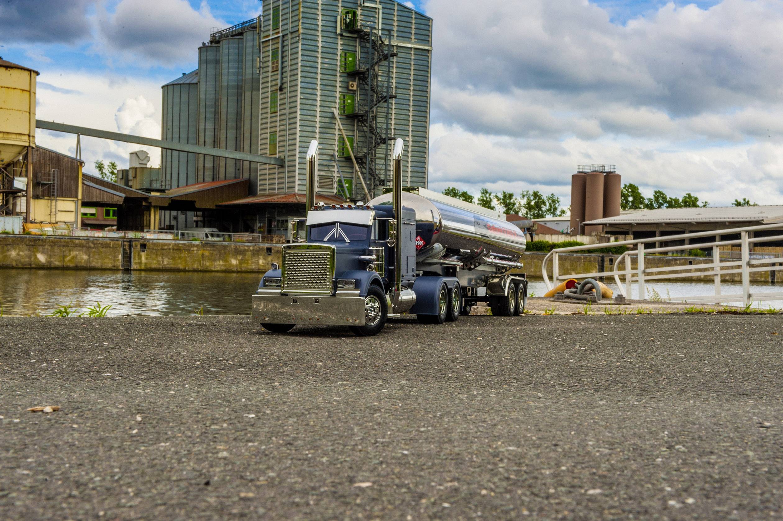 'Ein großer Lkw mit silbernem Tankanhänger steht auf einer Straße nahe einem Industriekomplex am Wasser unter blauem Himmel mit Wolken.'