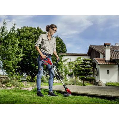 Eine Frau in Freizeitkleidung trimmt den Rasen neben einem Haus mit einem elektrischen Rasentrimmer. Im Hintergrund sind Bäume.