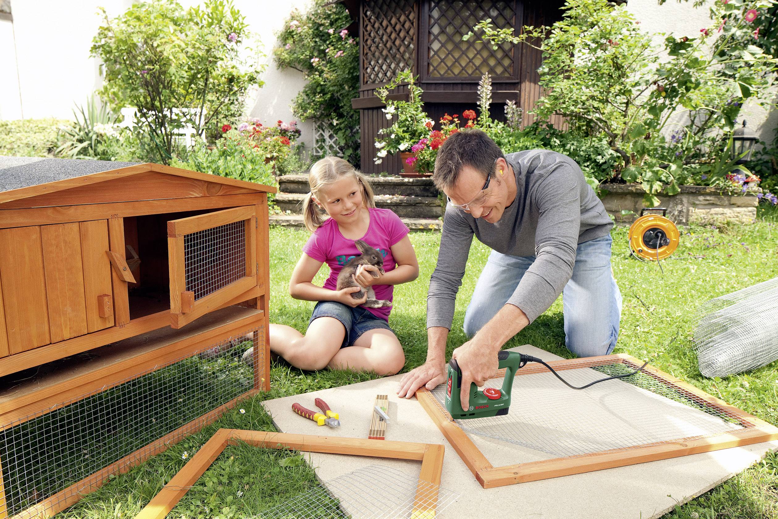 Ein Mann baut mit einem Mädchen einen Hasenkäfig im Garten. Der Mann verwendet ein Werkzeug, während das Mädchen ein Kaninchen hält.