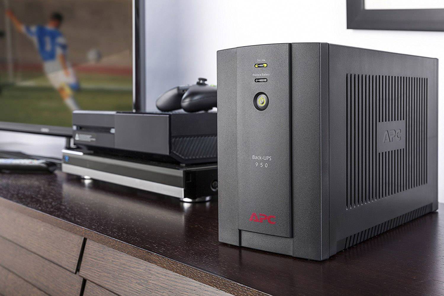Ein schwarzes APC-Backup-Gerät steht auf einem Holztisch neben einer Spielkonsole und einem Fernseher, der ein Fußballspiel zeigt.