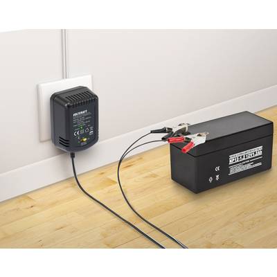 Ein schwarzes Batterieladegerät ist an eine Steckdose angeschlossen und lädt eine schwarze 12V-Batterie über Klemmen auf einem Holzboden.