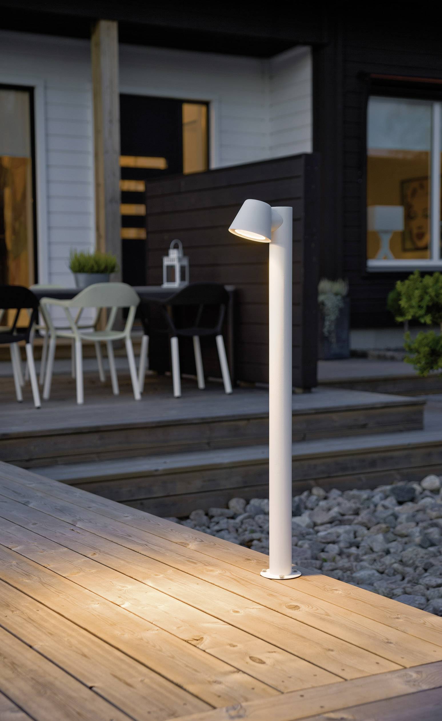 Weiße Stehlampe beleuchtet Holzterrasse neben Steinhaus. Im Hintergrund sind ein Tisch und Stühle auf einem erhöhten Deck sichtbar.