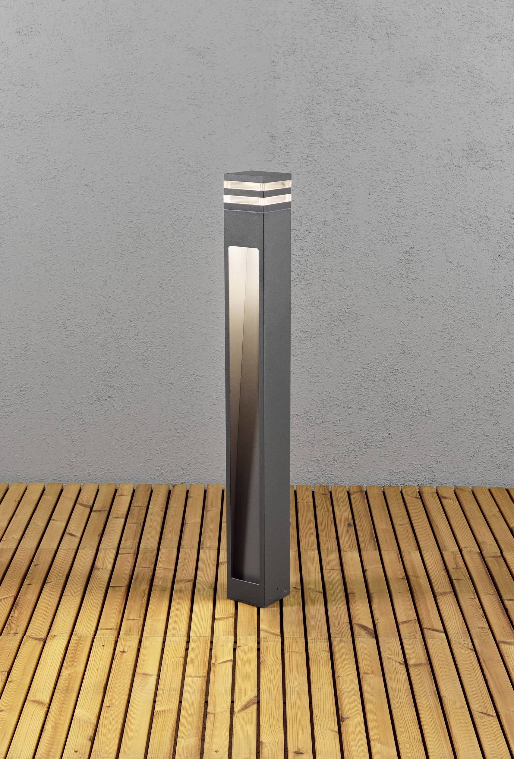 Ein moderner, schlanker Lichtpfosten aus Metall steht auf einem Holzdeck vor einer grauen Wand.