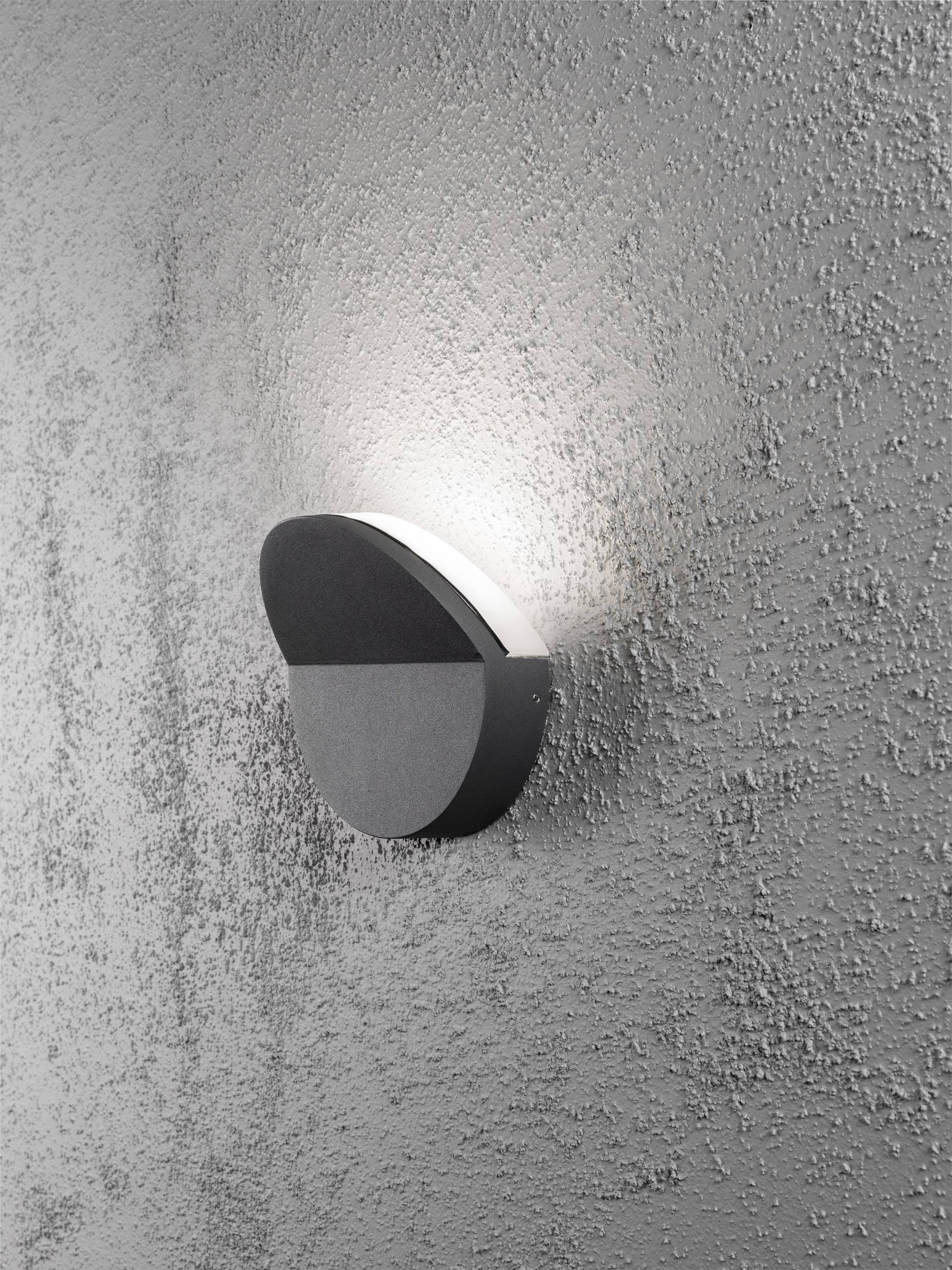 Wandleuchte mit rundem Gehäuse an einer strukturierten grauen Wand. Das Licht strahlt nach oben und erzeugt einen sanften Schein.
