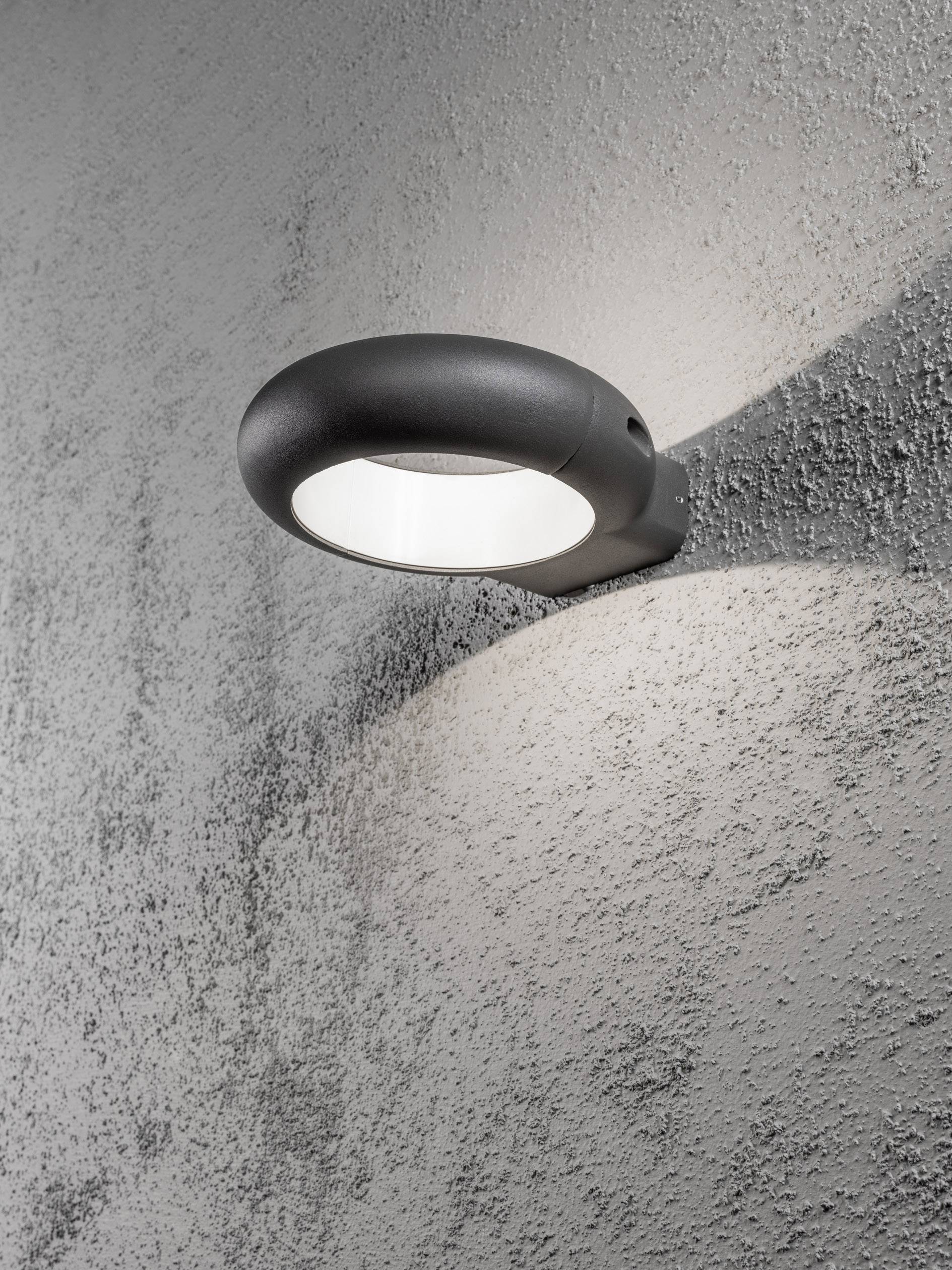 Eine moderne Wandlampe mit rundem, schwarzem Gehäuse strahlt warmes Licht auf eine strukturierte graue Betonwand.