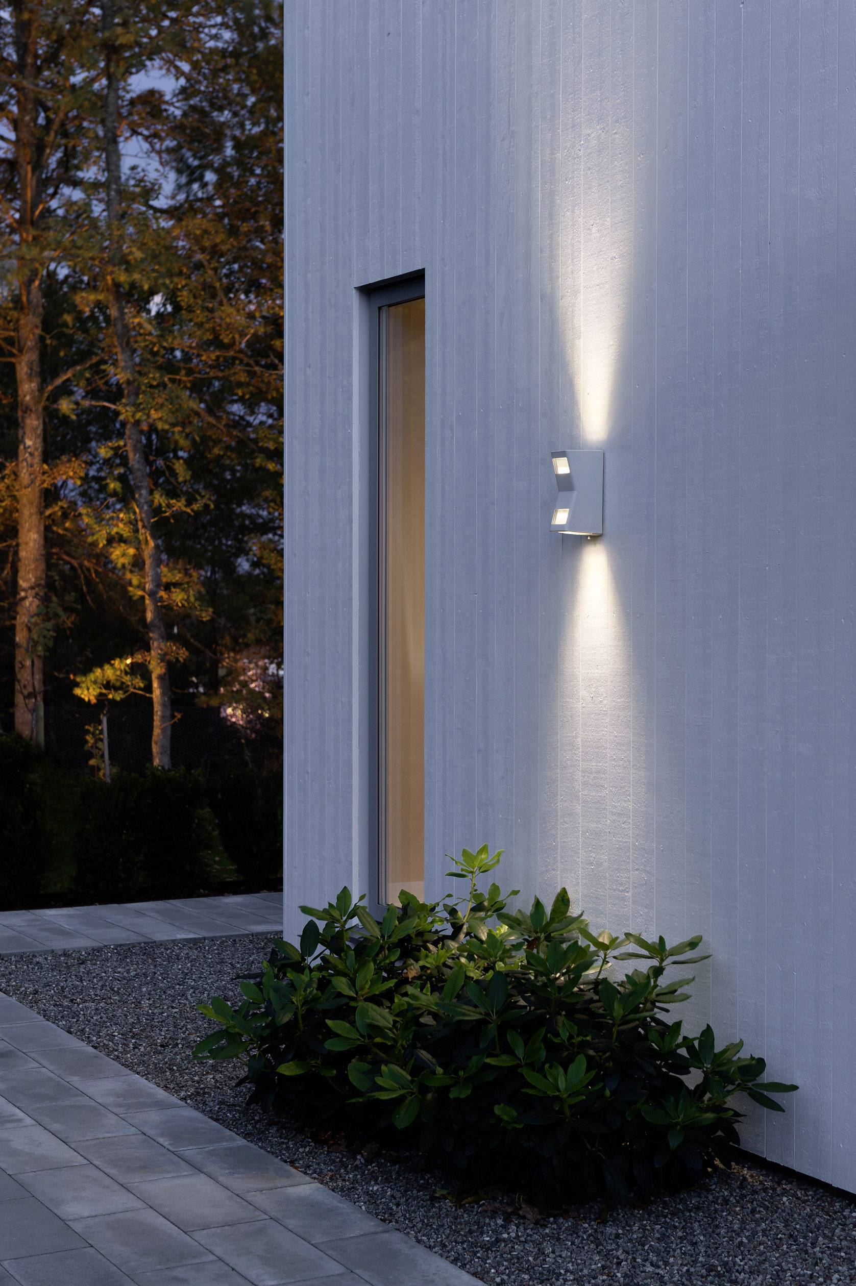 Außenaufnahme einer modernen Hausfassade mit Wandbeleuchtung, die Lichtstrahlen nach oben und unten wirft; im Vordergrund Pflanzen.