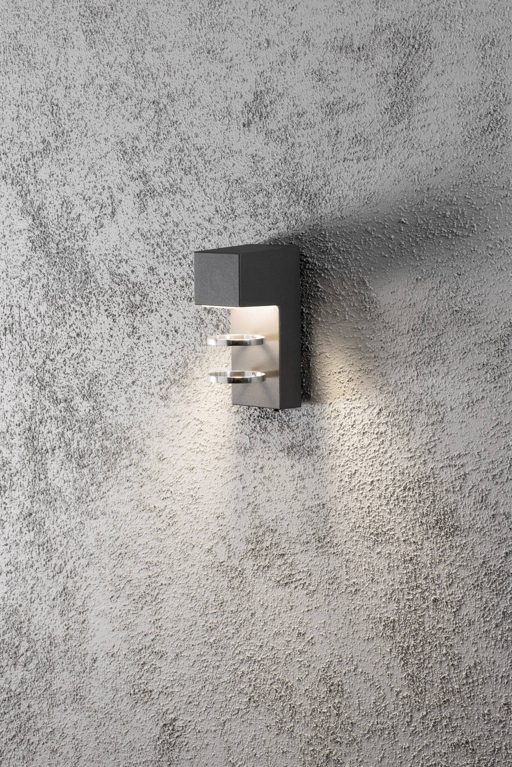 Eine moderne Wandleuchte mit rechteckigem Gehäuse strahlt warmes Licht an eine raue graue Wand.