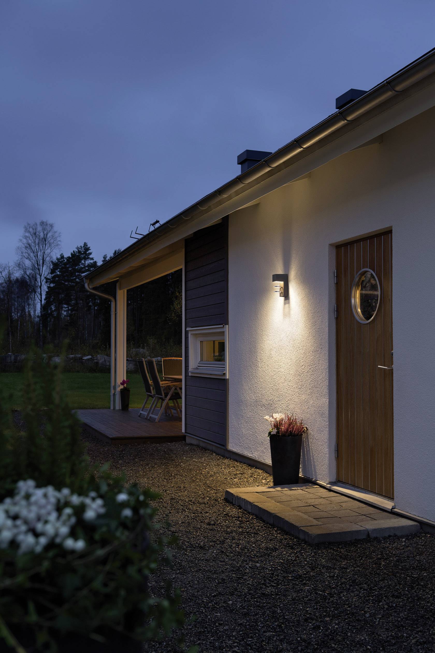 Ein Haus bei Dämmerung mit beleuchteter Außentür und Fenster. Auf der Veranda stehen Stühle. Im Vordergrund blühen Pflanzen.