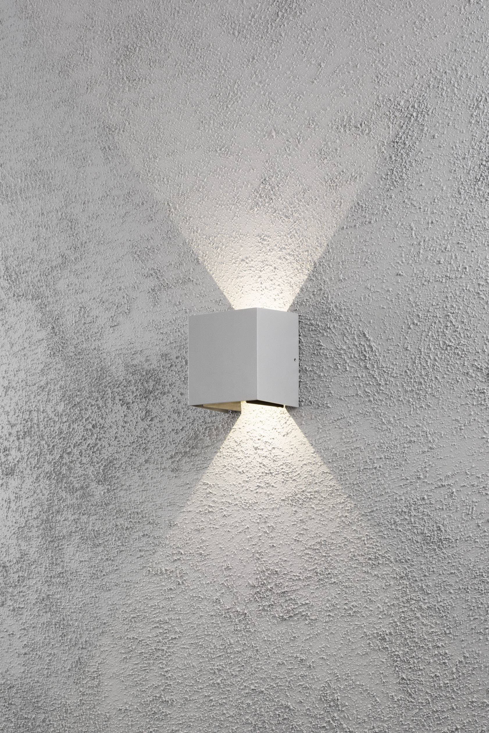 Wandleuchte mit quadratischem Gehäuse strahlt Licht nach oben und unten auf grob strukturierte, graue Wand. Minimalistisches Design.