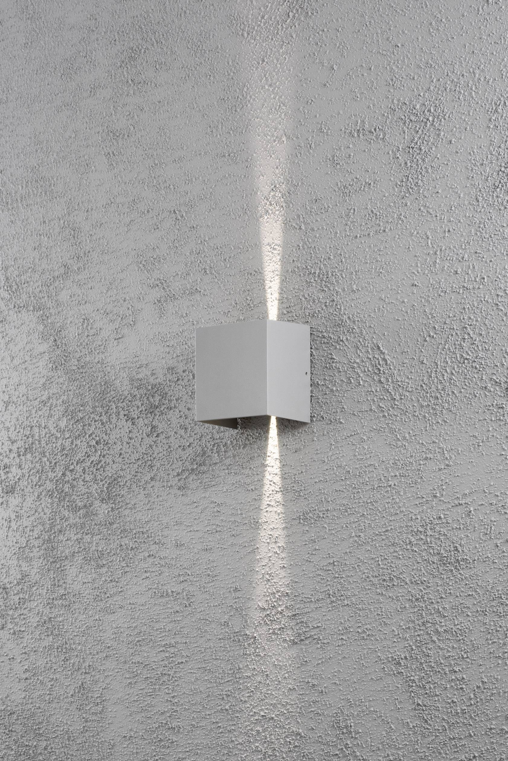 Eine graue, quadratische Wandleuchte auf einer rauen, grauen Wand, wirft Licht nach oben und unten in schmalen Kegeln.