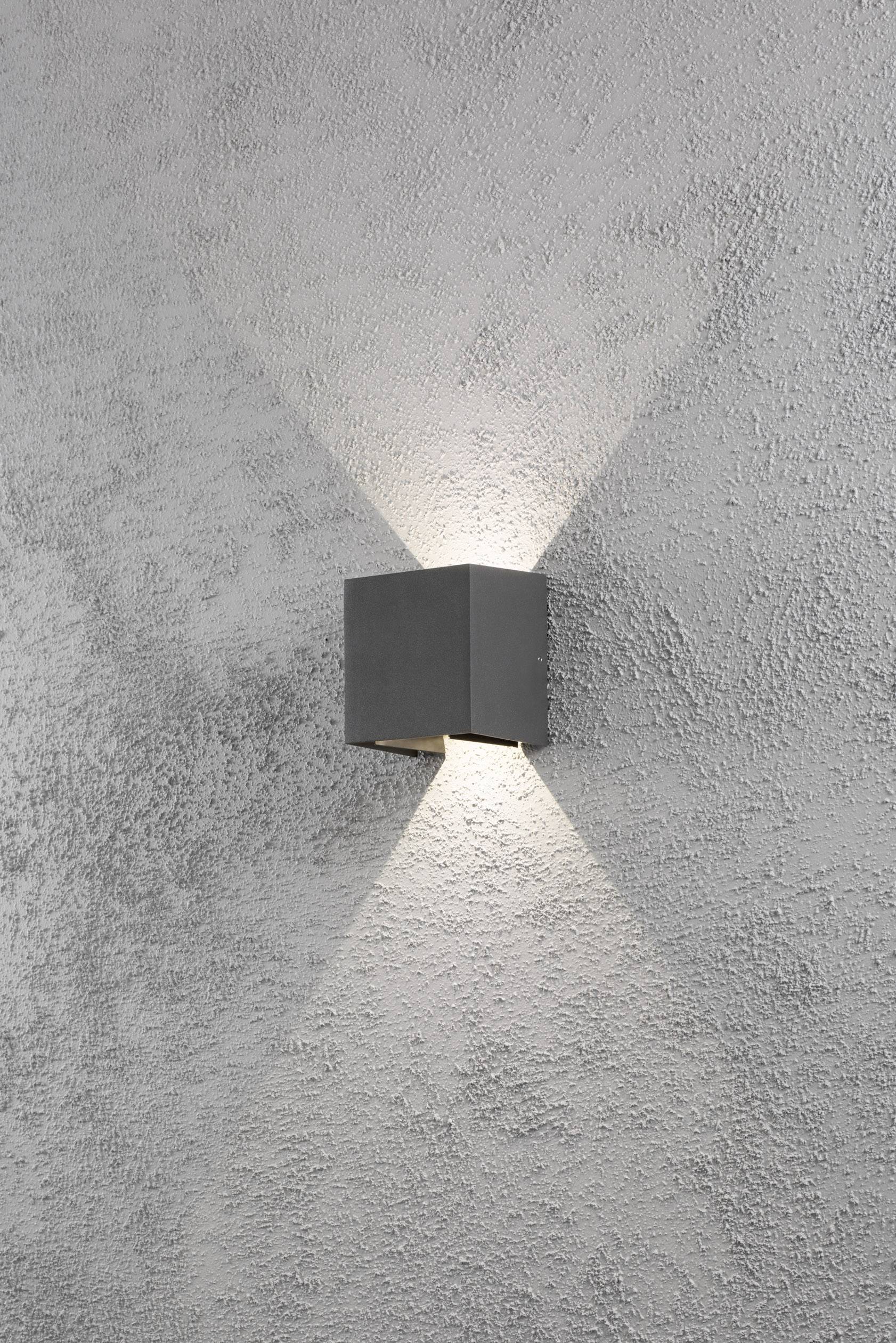Eine graue Wandlampe projiziert ein X-förmiges Lichtmuster auf eine grob strukturierte graue Wand.