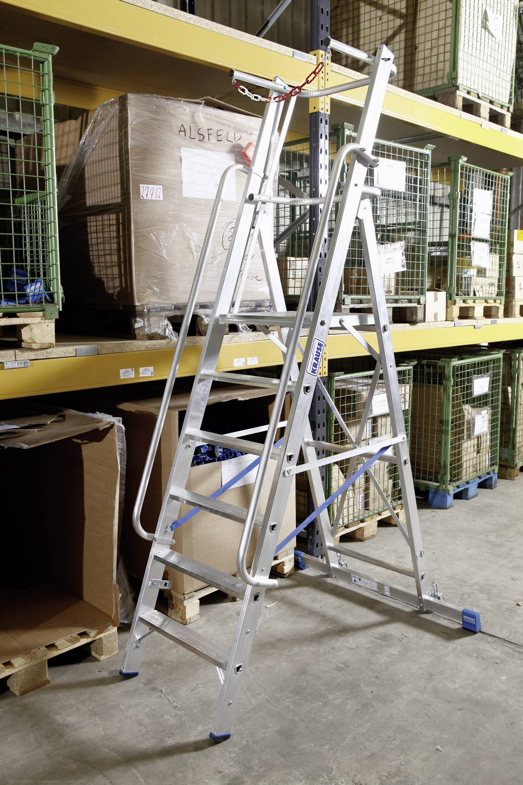 Eine bewegliche Aluminiumleiter steht in einem Lager zwischen Paletten und regalen. Sie hat Sicherheitsbügel und blauen Streben.