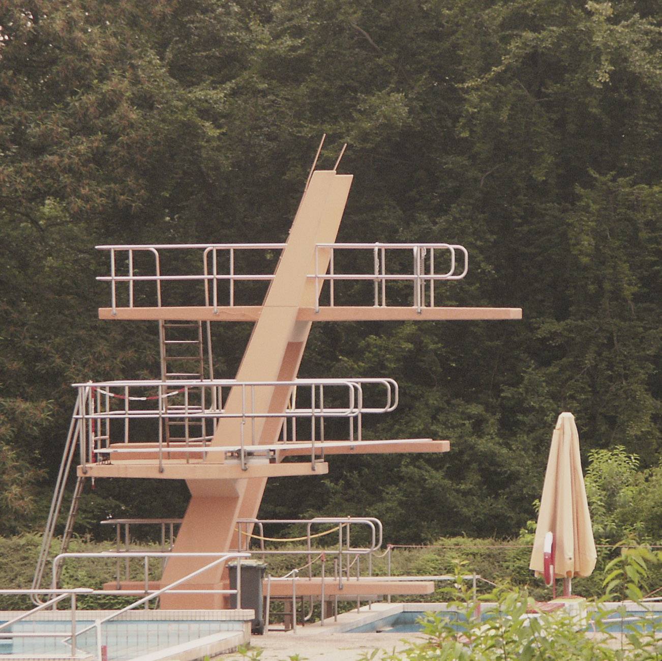 'Leeres Schwimmbecken mit einem dreistufigen Sprungturm im Freien. Umgeben von Bäumen und einem geschlossenen Sonnenschirm.'