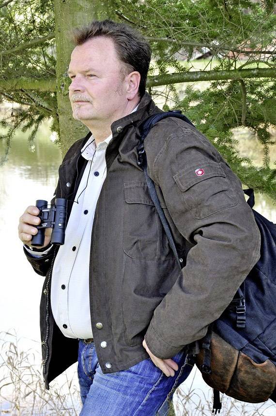 Eine Person in Freizeitkleidung steht im Freien, hält ein Fernglas und trägt einen Rucksack. Im Hintergrund sind Bäume und Wasser.