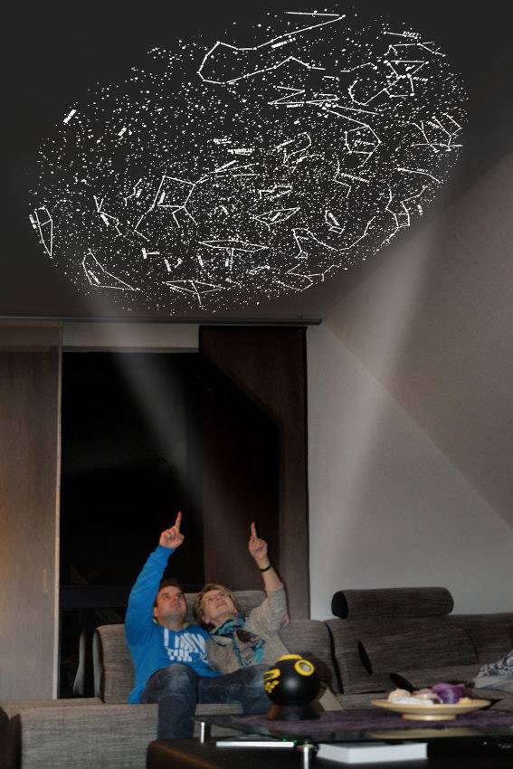Zwei Personen sitzen auf einem Sofa und zeigen auf eine projizierte Sternenkarte an der Zimmerdecke, die verschiedene Sternbilder darstellt.