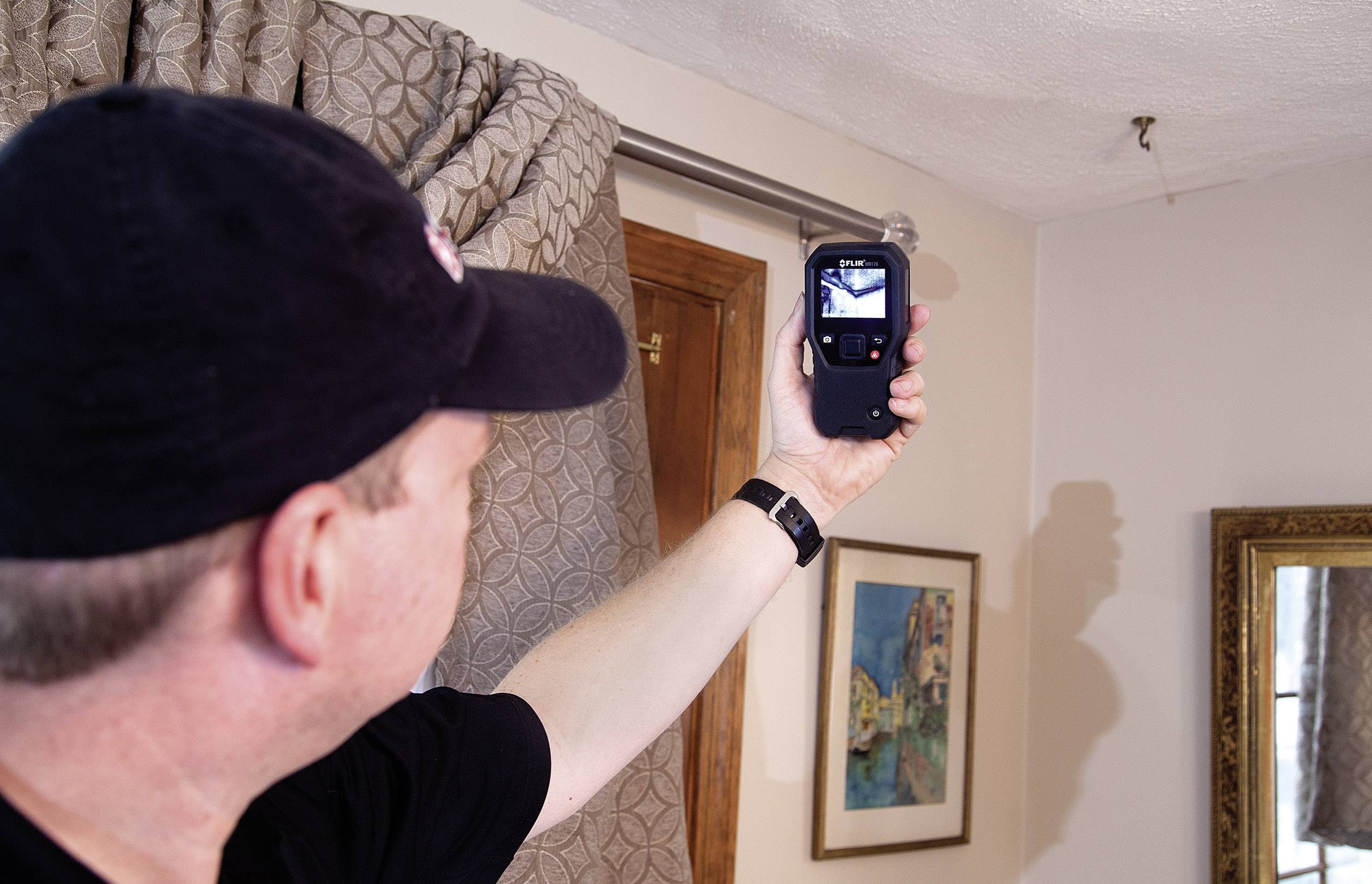 Eine Person fotografiert mit einem kleinen Gerät eine Wand im Raum. Im Hintergrund sind ein Fenster mit Vorhang und ein Bild zu sehen.