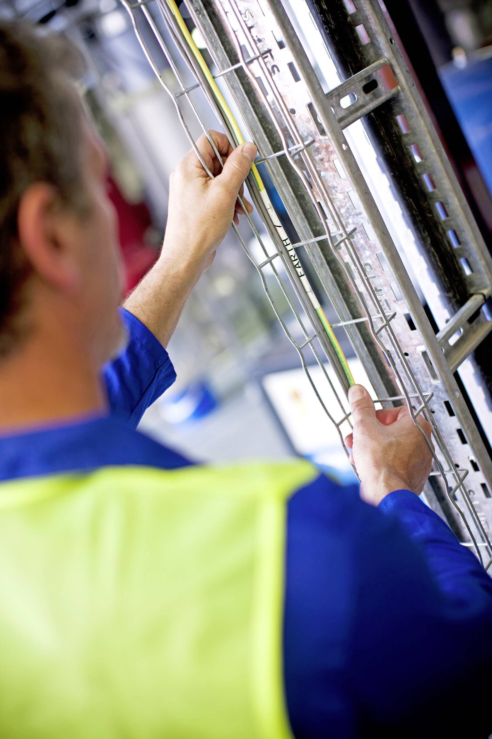 Mann in Arbeitskleidung misst mit einem Maßband ein Metallgitter. Im Hintergrund sind unscharfe industrielle Elemente zu sehen.