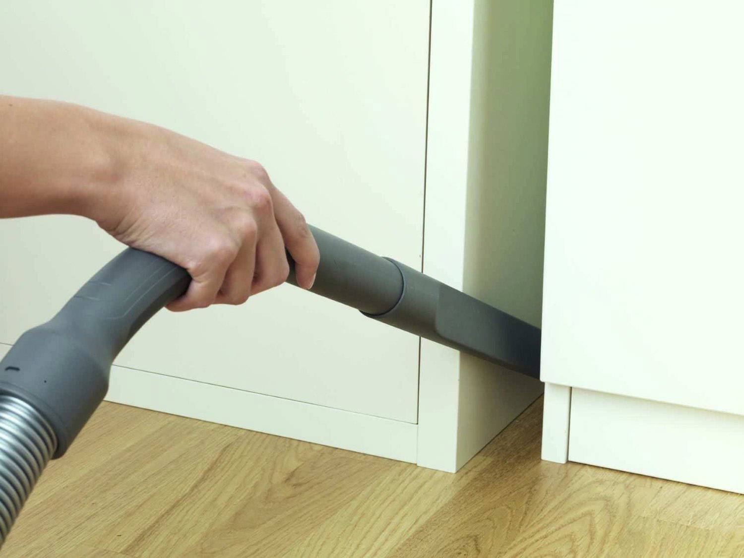 Eine Hand hält einen Staubsaugerschlauch, der Staub zwischen einem Schrank und dem Boden saugt. Der Boden scheint aus Holz zu sein.