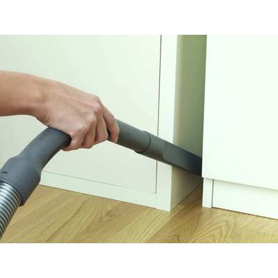 Eine Hand hält einen Staubsaugerschlauch, der Staub zwischen einem Schrank und dem Boden saugt. Der Boden scheint aus Holz zu sein.