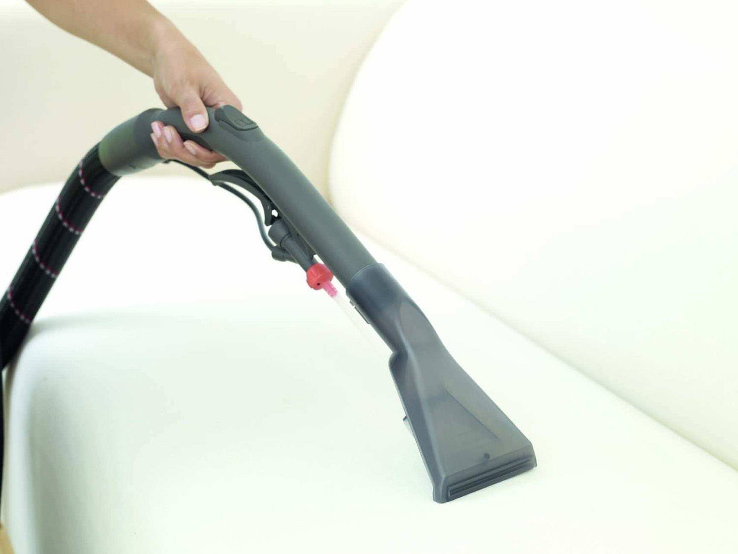 Eine Person verwendet einen Dampfreiniger, um ein helles Sofa zu reinigen. Der Dampfreiniger hat einen breiten schwarzen Aufsatz.