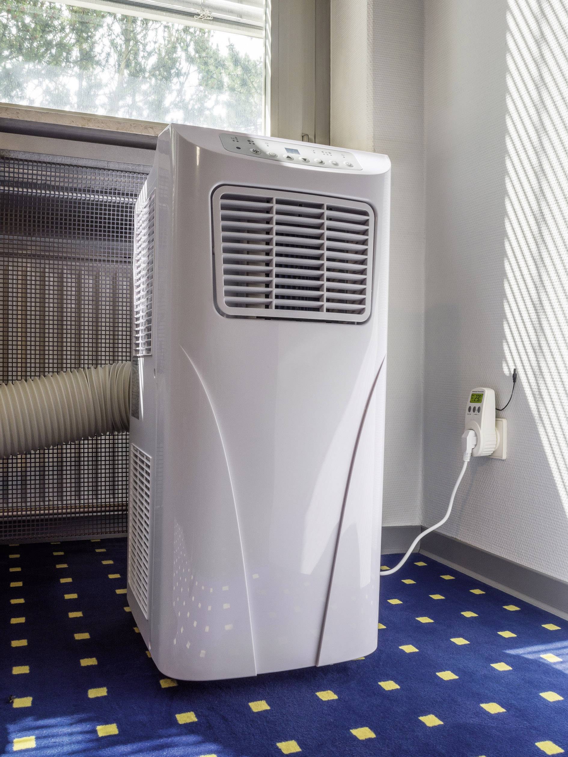 Eine mobile Klimaanlage steht in einem Raum auf einem blau-gelben Teppich. Sie ist über ein Rohr mit einem Fenster verbunden.
