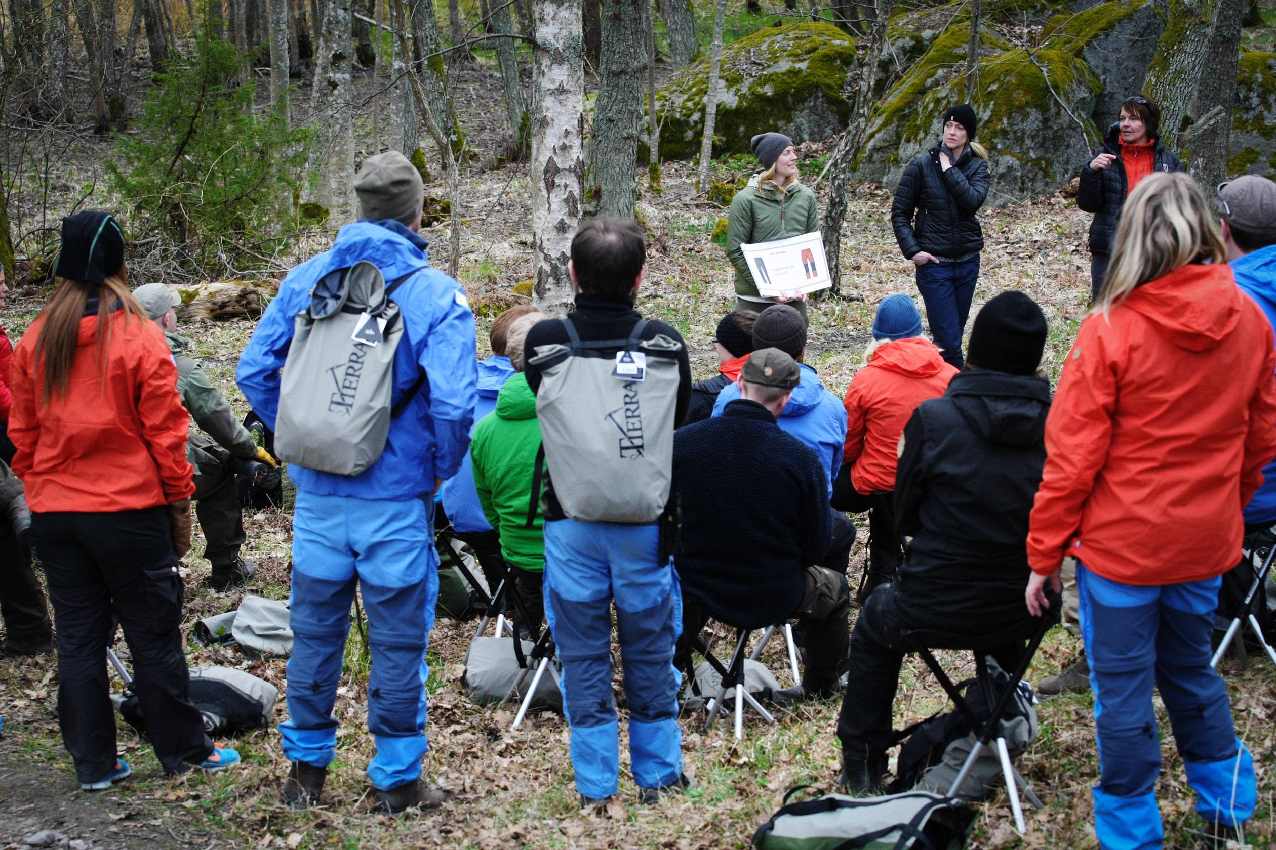 Eine Gruppe von Menschen sitzt im Wald auf Klappstühlen und hört einer Frau zu, die ein Bild präsentiert. Sie tragen Outdoor-Kleidung.