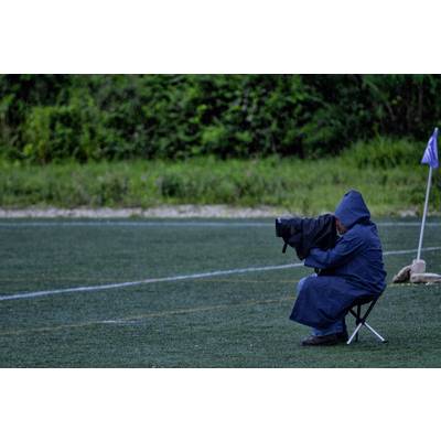 Person in dunklem Regenmantel sitzt am Spielfeldrand auf einem Klappstuhl und fotografiert ein Fußballspiel. Am Spielfeldrand eine Eckfahne.