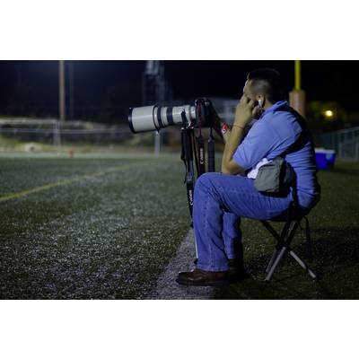 Ein Fotograf sitzt auf einem Stuhl am Spielfeldrand mit einer großen Kamera und Objektiv, konzentriert auf das Geschehen.