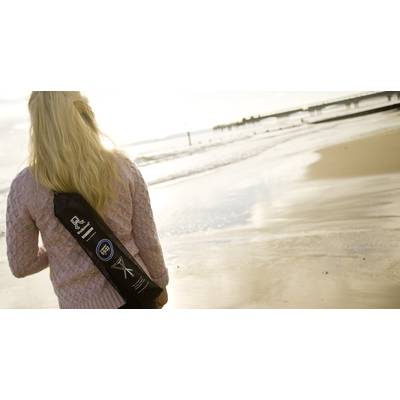 Eine Person geht am Strand entlang und trägt eine schwarze Tragetasche mit Logos. Der Himmel ist bewölkt, das Meer ist ruhig.