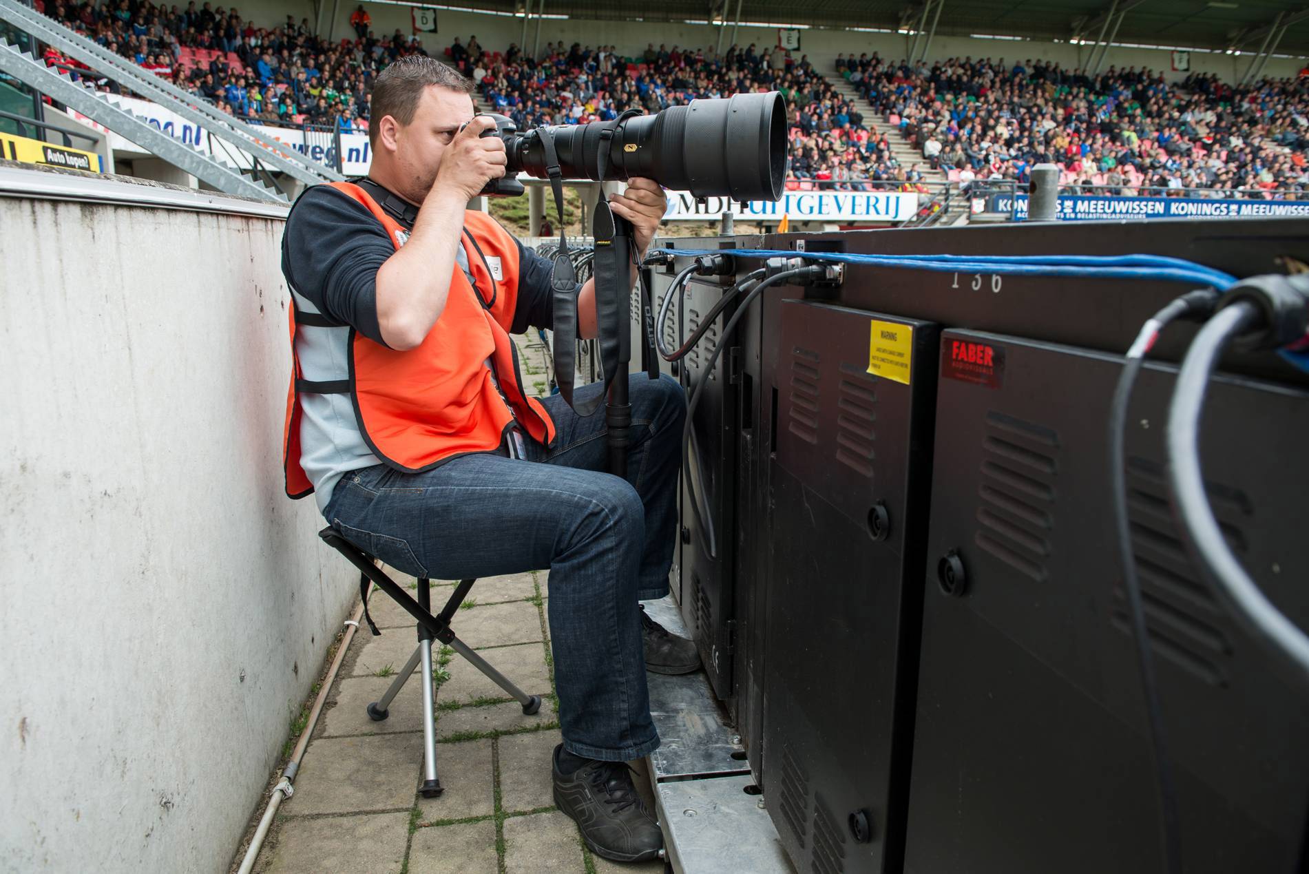 Ein Fotograf im Sportstadion sitzt neben technischen Geräten und hält eine große Kamera, um auf das Spielfeld zu fokussieren. Im Hintergrund Zuschauer auf Tribünen.