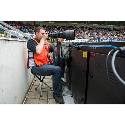 Ein Fotograf im Sportstadion sitzt neben technischen Geräten und hält eine große Kamera, um auf das Spielfeld zu fokussieren. Im Hintergrund Zuschauer auf Tribünen.