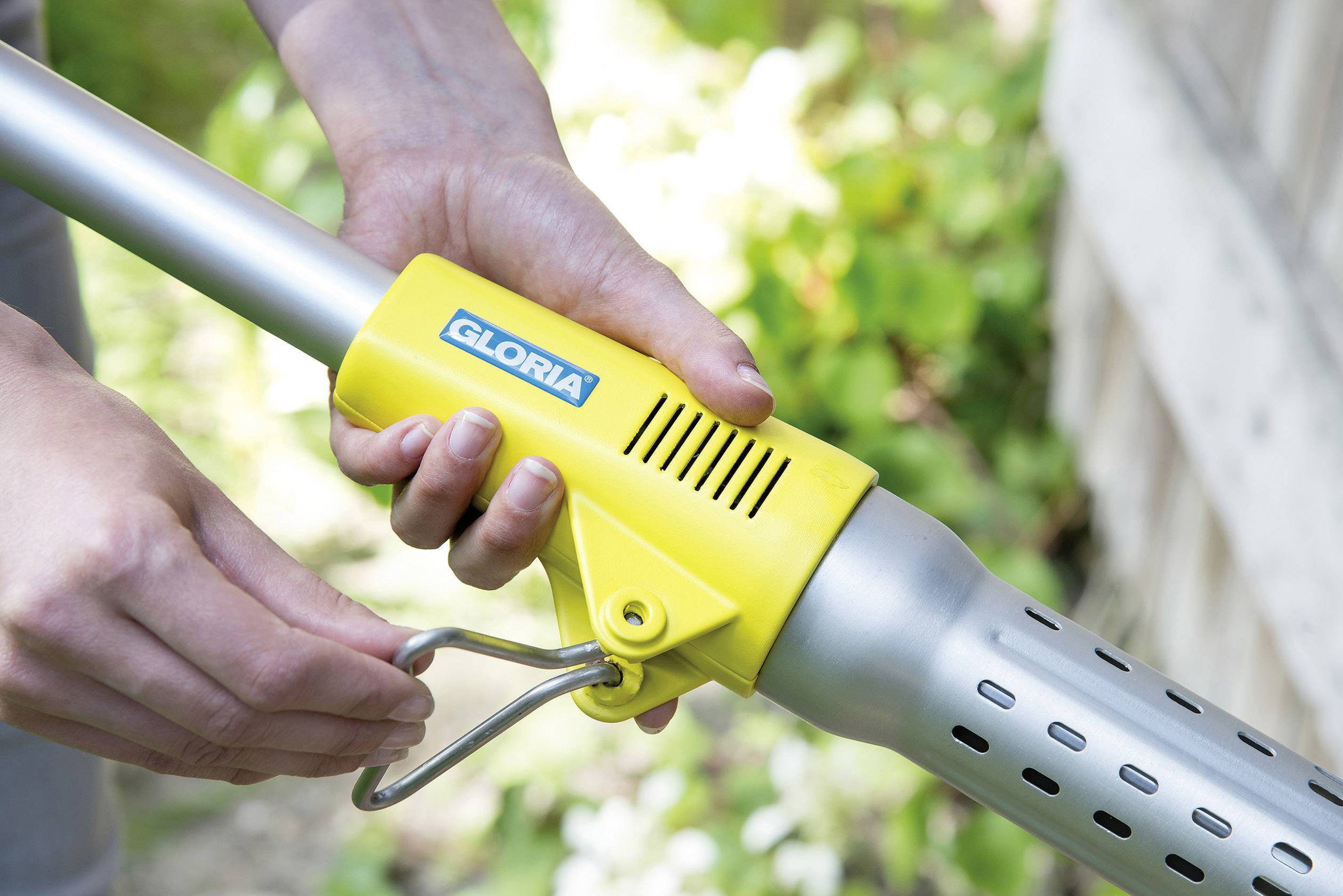 Eine Person hält ein gelbes Gartenwerkzeug mit Metallgriff und Lüftungsschlitzen. Das Werkzeug hat einen Aufdruck 'Gloria'.