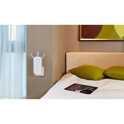 Ein WLAN-Repeater ist an der Wand eines Schlafzimmers neben einem Bett mit grünen Kissen installiert. Ein Tablet liegt auf dem Bett.