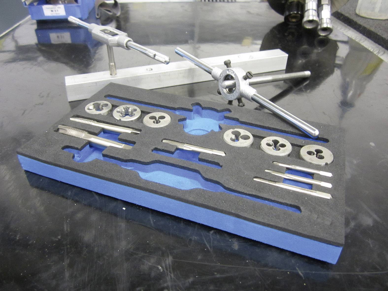 Eine Auswahl von Metallgewindeschneidwerkzeugen auf einem Arbeitstisch, darunter Schneideisen und Halterungen, geordnet in einem Schaumstoffhalter.