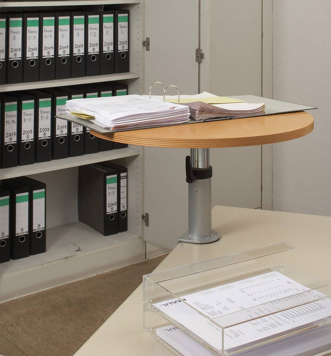 Ein Büro mit Ordnern im Regal und einem Stapel Papier auf einem runden Tisch. Im Vordergrund befindet sich ein Papierfach mit Dokumenten.