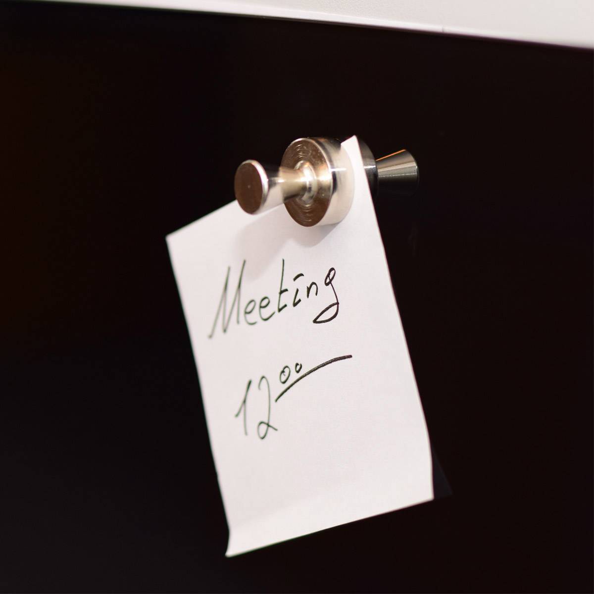Ein Zettel mit der handgeschriebenen Notiz 'Meeting 12:00' ist an einer schwarzen Tafel mit einem Magnet befestigt.