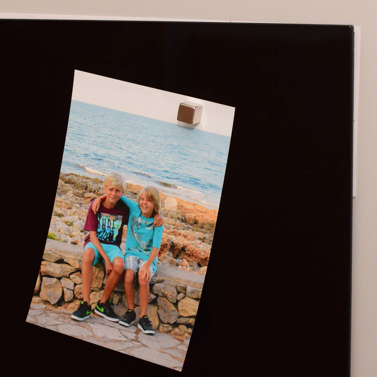 Zwei Kinder sitzen auf einer Steinmauer am Meer. Das Bild ist schräg an einer schwarzen Fläche angebracht. Das Wetter scheint sonnig zu sein.