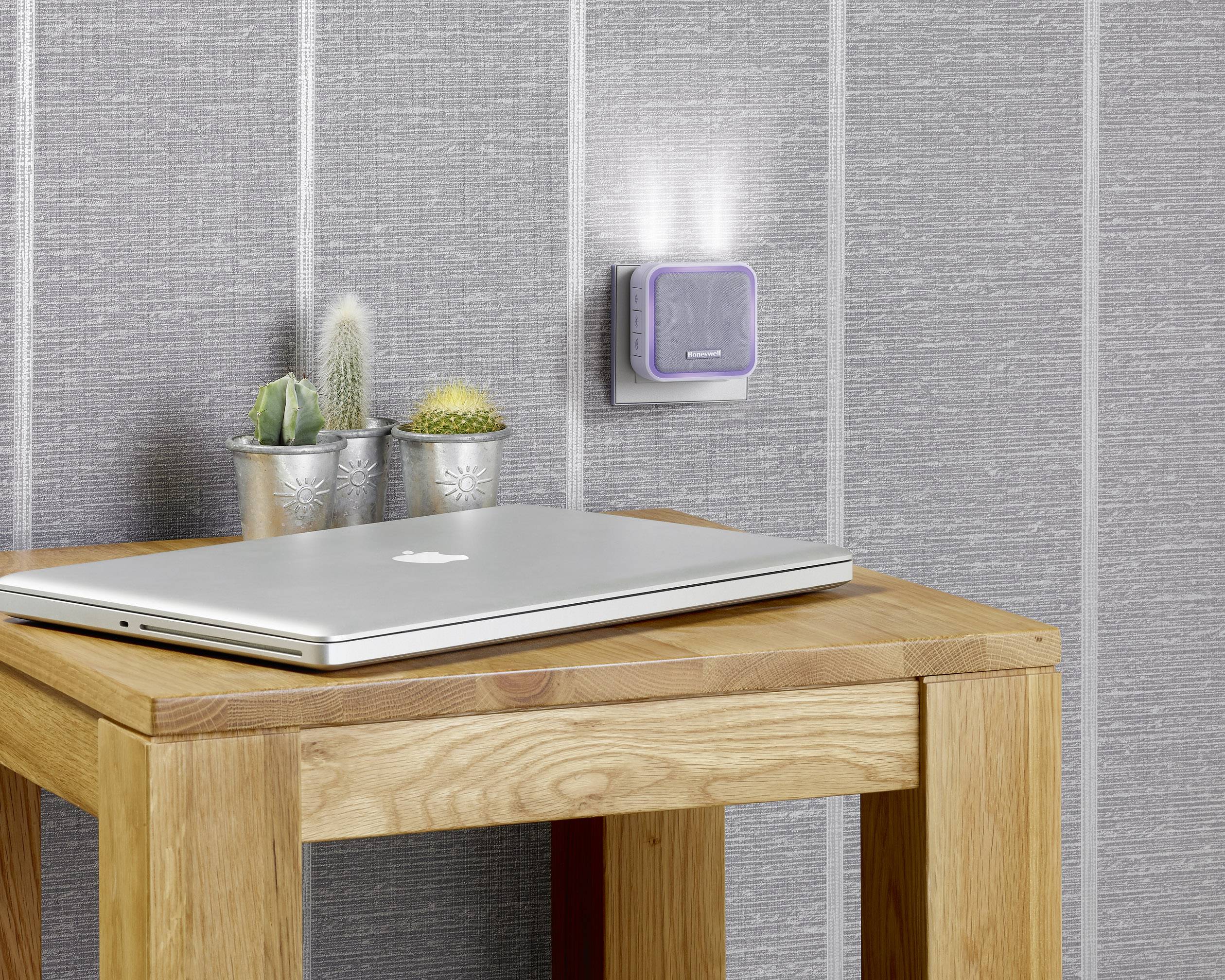 Ein Holz-Tisch mit einem geschlossenen Laptop und drei kleinen Kakteen darauf. An der grauen Wand im Hintergrund ist ein Nachtlicht befestigt.