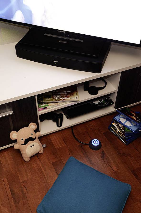 Ein Wohnzimmerregal mit Büchern und Elektronik. Ein Fernseher steht oben, darunter Konsolen-Zubehör. Ein Plüschbär mit Schnurrbart ist links.