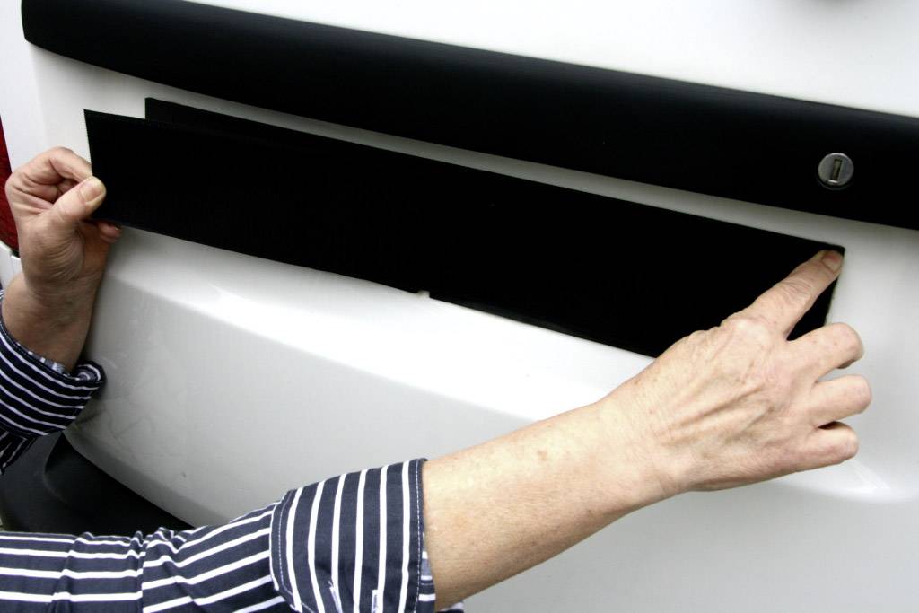 Eine Person befestigt ein schwarzes Schild an einem weißen Fahrzeug, möglicherweise ein Kennzeichen- oder Einparkschutz.