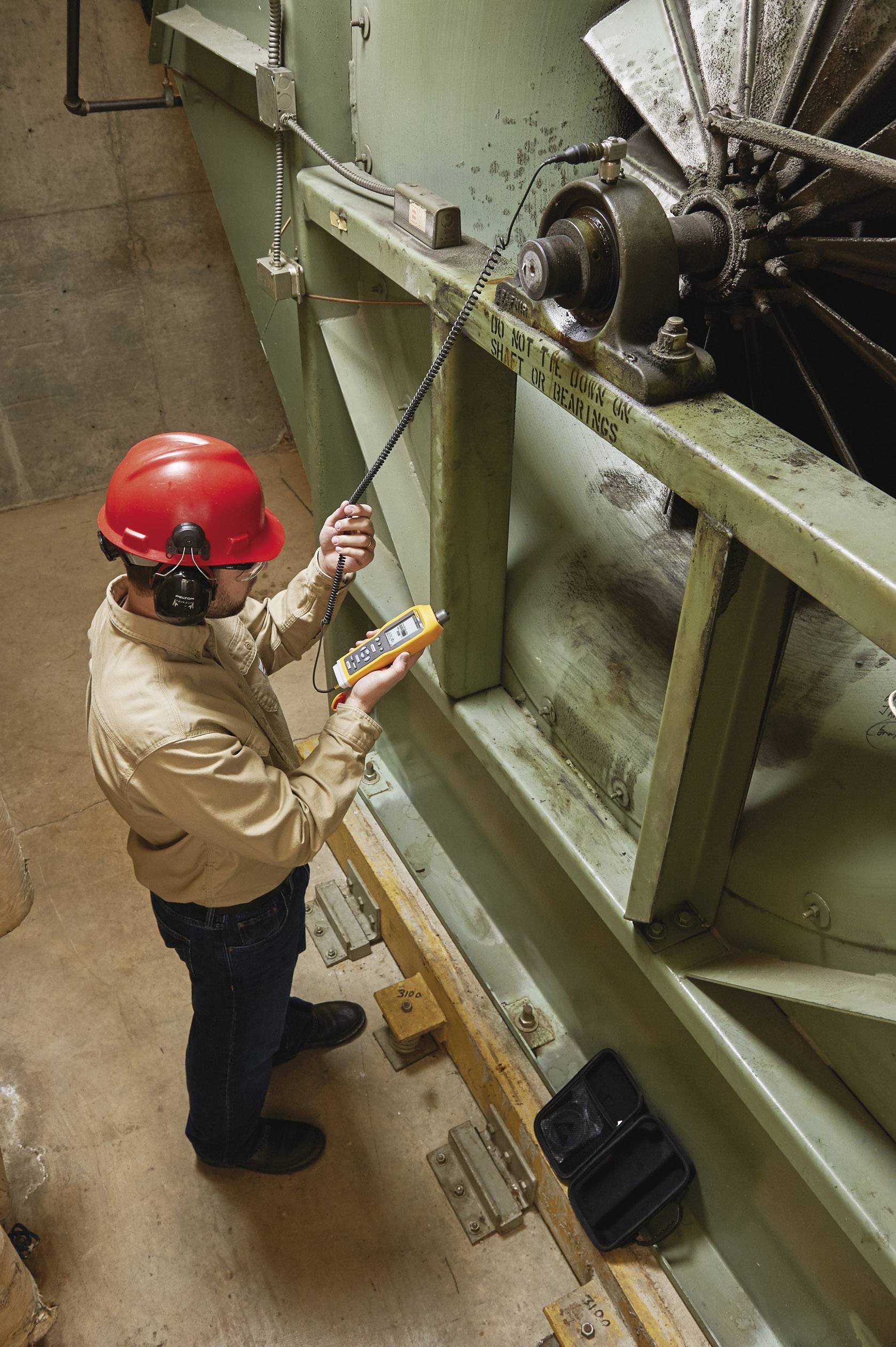 Ein Techniker mit rotem Helm misst mit einem Gerät die Schwingungen einer großen industriellen Maschine.