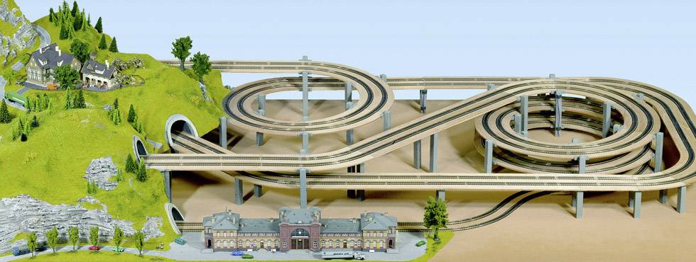 Eine komplexe Modelleisenbahnlandschaft mit mehrstöckigen Schienen und einem Bahnhof im Vordergrund. Links sind Hügel mit Tunneln.