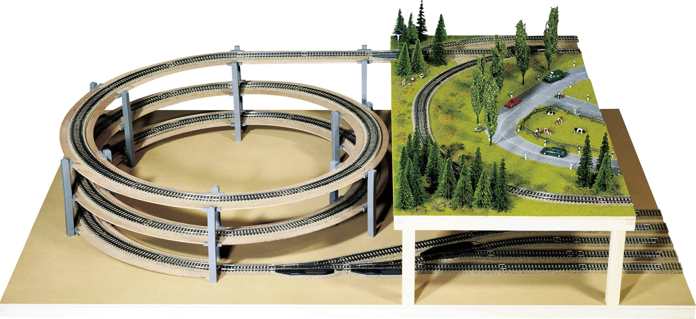 Modelleisenbahn mit mehrstöckigem Gleisaufbau links und landschaftlicher Szene rechts mit Bäumen, Autos und Figuren.