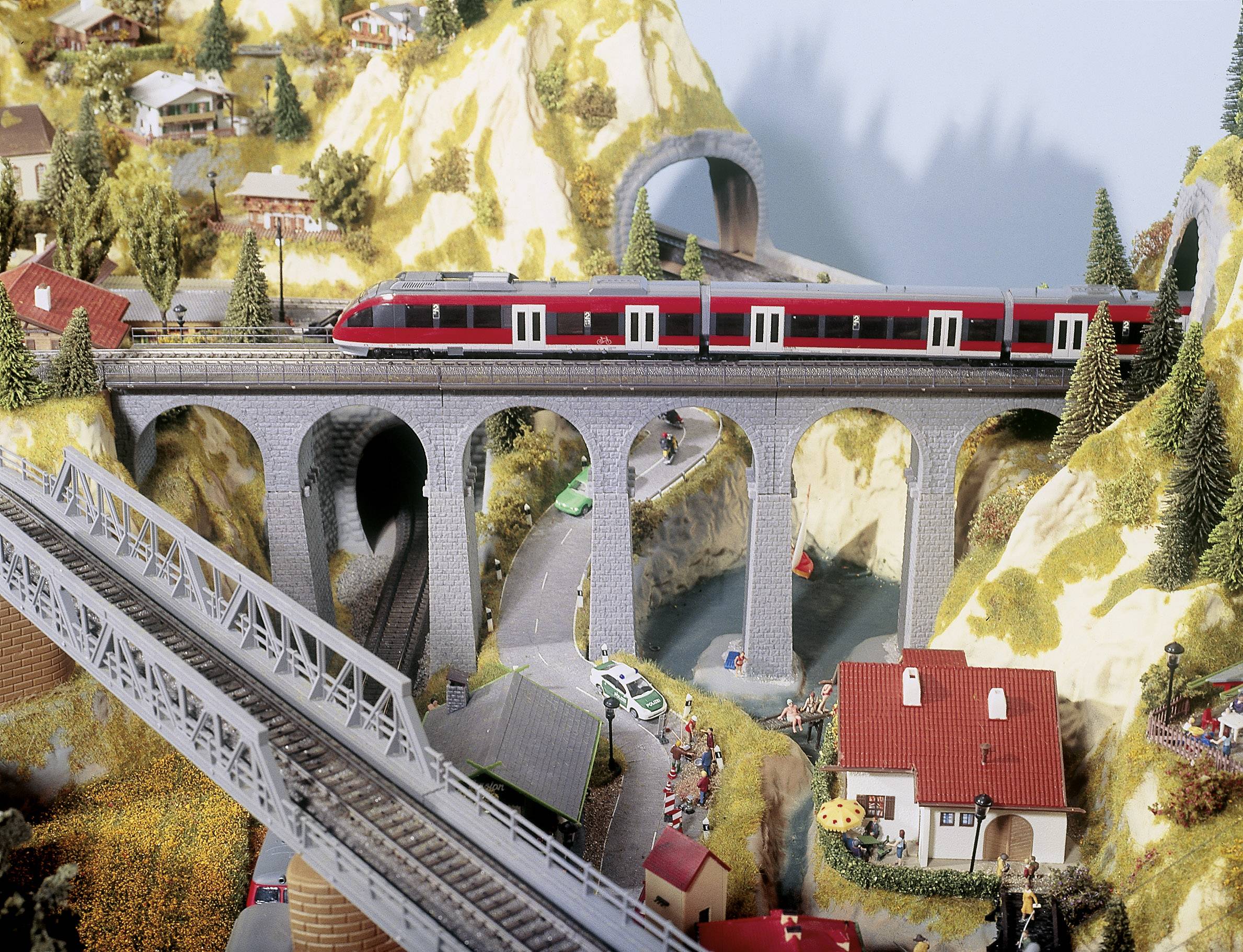 Eine Modelleisenbahn fährt auf einem Viadukt durch eine bergige Landschaft mit Tunneln und Gebäuden, die eine Miniaturwelt darstellen.