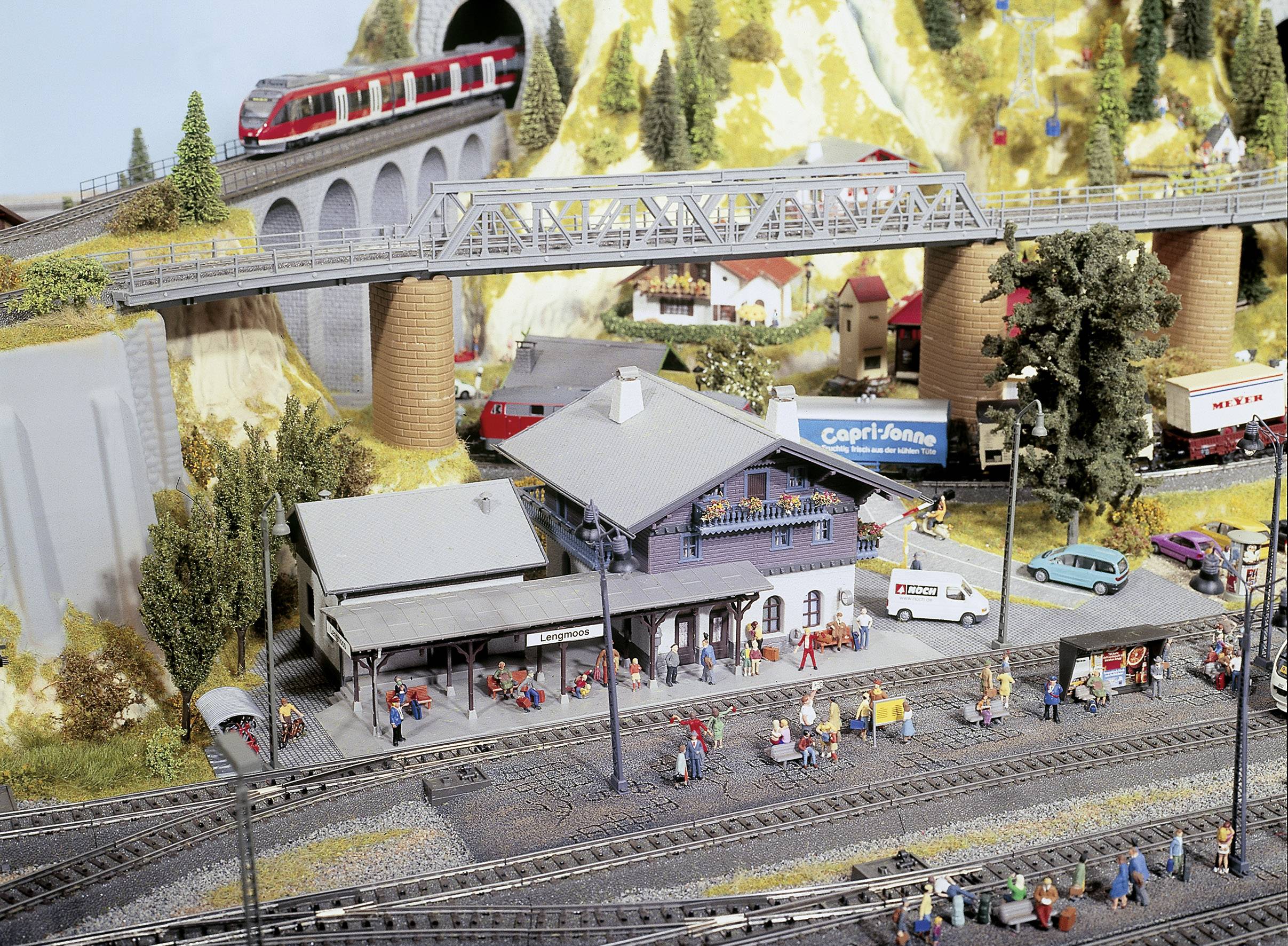 Modelleisenbahnlandschaft mit Bahnhof, Zügen auf Brücke, Autos und Miniaturfiguren. Detailreiche Darstellung einer Stadt.
