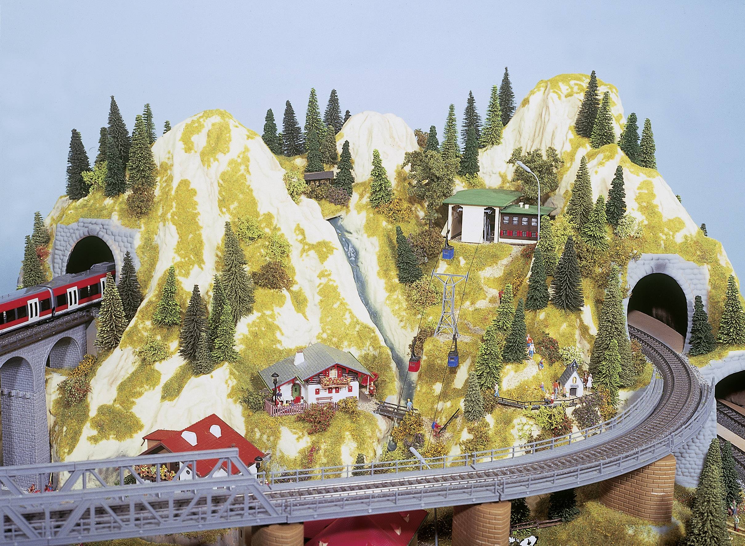 Eisenbahnmodell mit Zug, der über Brücke durch eine bergige, mit Bäumen bewachsene Landschaft fährt; zeigt kleine Gebäude und Seilbahn.