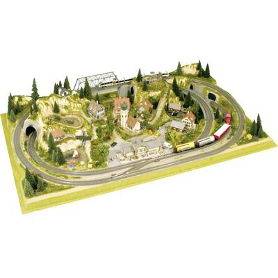 Eine Modellbahnanlage zeigt eine Landschaft mit Hügeln, Brücken, Häusern und Tunneln. Mehrere Züge fahren auf den Gleisen.