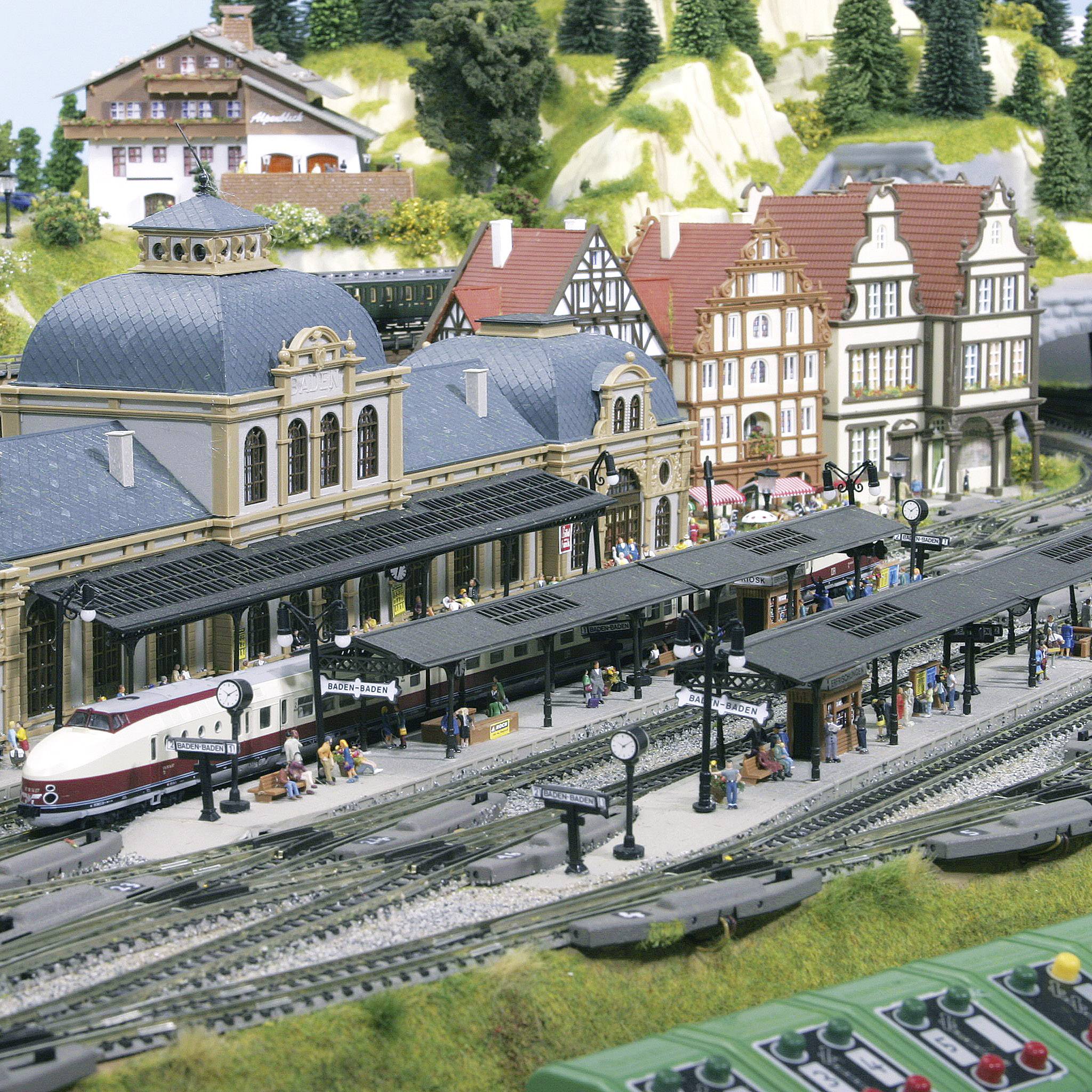Modelleisenbahn mit Bahnhofsszene. Mehrere Züge auf den Gleisen, umgeben von Gebäuden und kleinen Figuren, die den Bahnhof beleben.